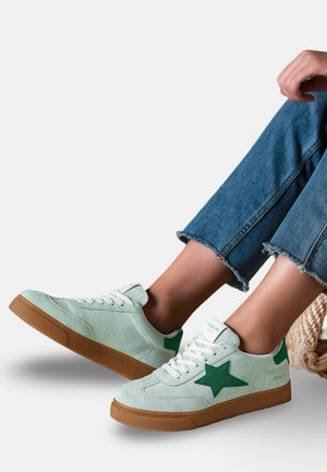 Pieds chaussés de baskets en suède vert clair avec des logos d'étoile verts, des lacets blancs et des semelles en gomme, assorties à un jean bleu à ourlet effiloché.
