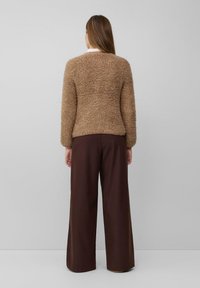 Vrouw met lang haar in een beige textuurjack en brede bruine broek, staand tegen een effen lichtgrijze achtergrond, met de rug naar de camera gekeerd.