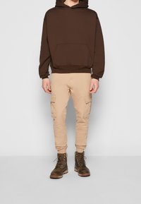 Sweat-shirt marron à capuche avec une poche frontale, associé à un pantalon cargo beige avec des poches latérales, et des bottes marron à lacets.