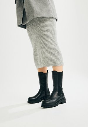 Personne portant une jupe en maille côtelée grise et des bottes en cuir noires mi-mollet avec des semelles épaisses sur un fond clair uni.