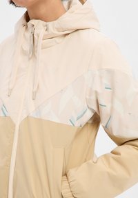 Veste coupe-vent beige avec une capuche, présentant une partie supérieure à motifs dans des couleurs claires, des manches avec poignet élastique, et une fermeture éclair à l'avant.