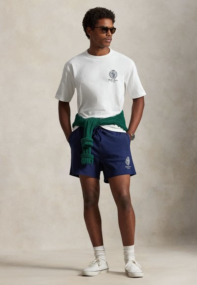 Λευκό βαμβακερό μπλουζάκι με τύπωμα λογότυπου, συνδυασμένο με σκούρες μπλε βερμούδες με κεντητό σχέδιο. Πράσινο πουλόβερ δεμένο στη μέση, λευκά αθλητικά παπούτσια.