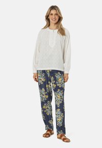 Blusa bianca a maniche lunghe con un motivo testurizzato e una patta con bottoni, abbinata a pantaloni blu con stampa floreale e sandali marroni.
