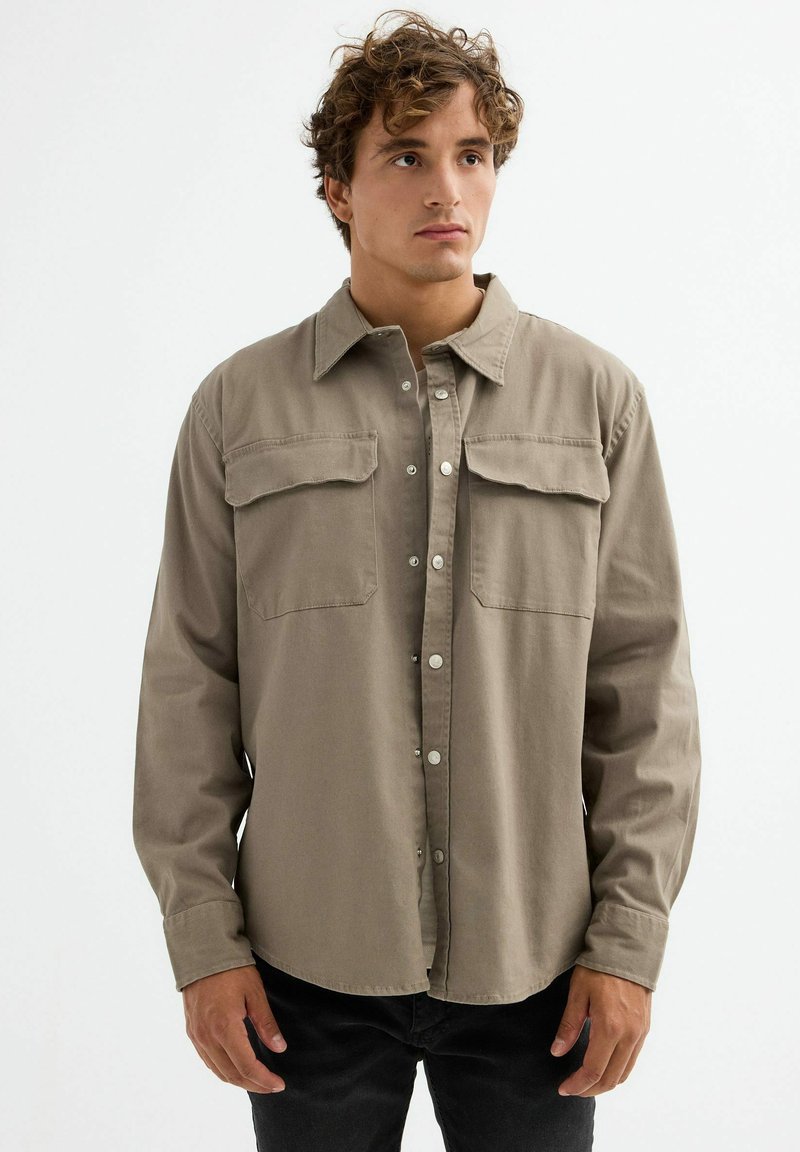 Camisa de manga larga beige, hecha de una tela suave, con dos bolsillos en el pecho, cierre de botones y un cuello clásico. Superficie texturizada, corte relajado.