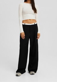 Schwarze weit geschnittene Hosen mit einem weißen Bund, kombiniert mit einem enganliegenden cremefarbenen Oberteil mit langen Ärmeln. Das Material wirkt weich und glatt.