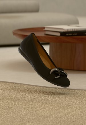 Schwarzer Ballerina mit glatter, lederähnlicher Oberfläche, runder Zehenpartie und einer metallischen Schleifenapplikation. Verfügt über eine profilierte Sohle für besseren Halt.