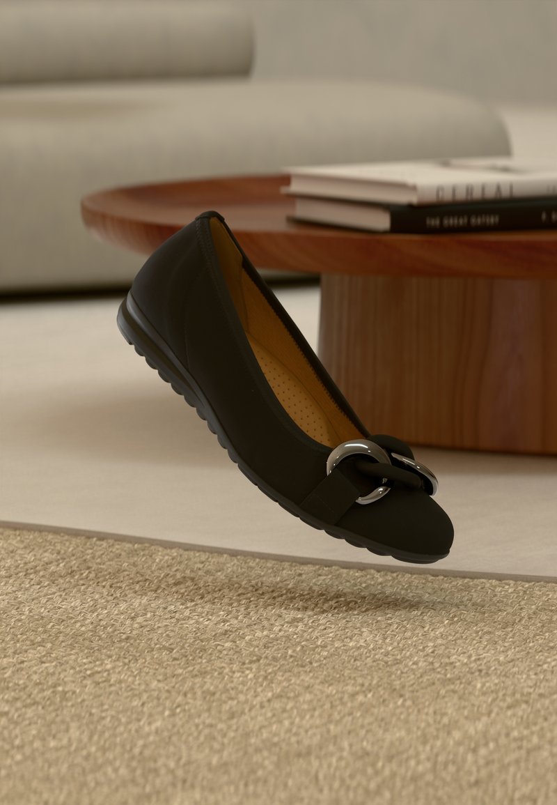 Svart ballettsko med glatt skinnlignende ytre, rund tå og en metallisk sløyfedetalj. Har en teksturert såle for grep.