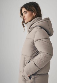 Beige Pufferjacke mit hohem Kragen, Kapuze, seitlichen Taschen und gesteppter Textur. Glattes Material mit sichtbaren Nähten und lässiger Passform.
