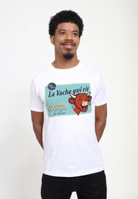 Vit t-shirt med en färgglad vintage-grafik av en leende ko med texten "La Vache qui rit" och "LA CRÈME DE GRUYÈRE de Luxe."