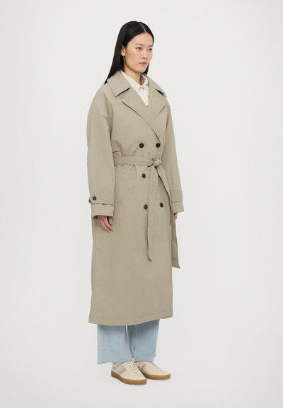 Beige trench coat with a double-breasted design, large collar, waist tie, and button accents. Worn over a light shirt and loose jeans.