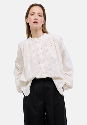 Femme portant un chemisier blanc ample à boutons avec des motifs ajourés verticaux et un pantalon ample noir à jambes larges, debout les mains dans les poches.