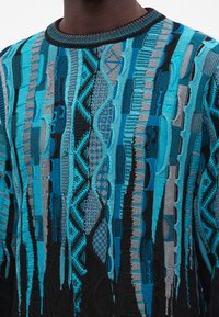 Gros plan d'un pull texturé avec des motifs verticaux dans des nuances de bleu, noir et gris sur une personne à la peau foncée.