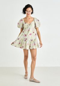 Rochie florală de un verde deschis, cu mâneci bufante, decolteu cu șnur și tiv cu volane în straturi. Purtați-o cu balerini transparenți.