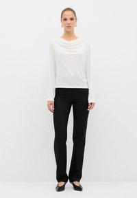 Witte longsleeve top met gedrapeerde halslijn, gecombineerd met zwarte rechte broek. Model draagt platte zwarte schoenen en grote gouden oorbellen.