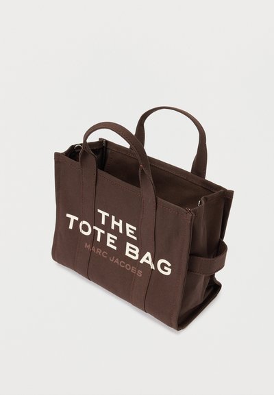 Καφέ καμβάς τσάντα tote με δύο λαβές και την επιγραφή "Η ΤΣΑΝΤΑ TOTE MARC JACOBS" τυπωμένη σε λευκό και ανοιχτό καφέ μπροστά.