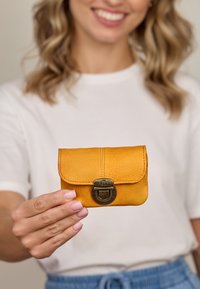 Geltonas odinis kortelių laikiklis su išlenktu dangčiu, tekstūruota paviršiumi ir sendintu žalvariniu užsegimu, laikomas rankoje prieš neutralų foną.