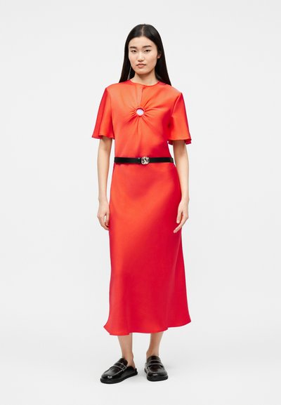 Γυναίκα που στέκεται με φωτεινό κόκκινο, μίντι φόρεμα με κοντά μανίκια, με κυκλικό άνοιγμα στο στήθος, μαύρη ζώνη και μαύρα παπούτσια τύπου slip-on.