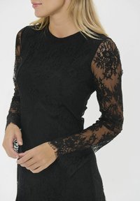 Juoda nėriniuota suknelė su ilgomis permatomomis nėrinių rankovėmis, gėlių raštais, prigludusia siluetu ir glotnia audinio kūnu.