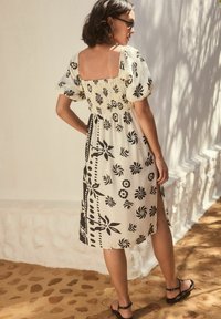 En knälång klänning med svart blommönster på en krämfärgad bakgrund, med smockat liv och puffärmar, slits på sidan och platta sandaler.