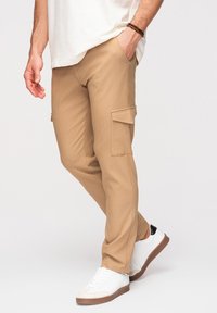 Homme portant un pantalon cargo beige, des baskets blanches avec des semelles marron, et une chemise blanche, une main dans la poche et un bracelet au poignet.