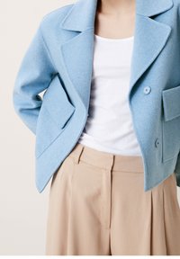 Ljusa blå yllejacka med snedskuren krage, två framfickor och två blå knappar. Matchad med krämfärgade plisserade byxor.