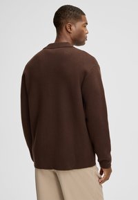 Brązowy sweterek robiony na drutach o teksturowanym wykończeniu, z kołnierzem i luźnym krojem, w połączeniu z jasnobeżowymi spodniami z prążkowanej tkaniny.