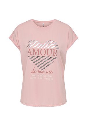 Joven mujer de pie con una camiseta rosa con diseño y texto, y pantalones cortos negros con borde en forma de concha, combinados con sandalias negras deslizantes.
