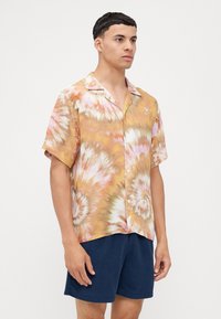 Camisa de botones de manga corta en patrón de teñido anudado en naranja y amarillo, hecha de una tela ligera; cuenta con un collar tipo camp y un corte relajado.