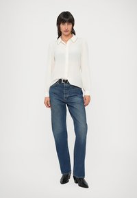Blouse blanche à manches longues avec une patte de boutonnage devant et un col texturé, associée à un jean bleu à jambes droites et des bottines noires.