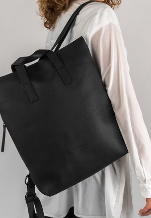 Henkilö, jolla on päällään valkoinen pitkähihainen paita ja joka kantaa suurta mustaa, kuvioitua nahkasta valmistettua olkalaukkua, jossa on kaksi kahvaa, olkapäällä.