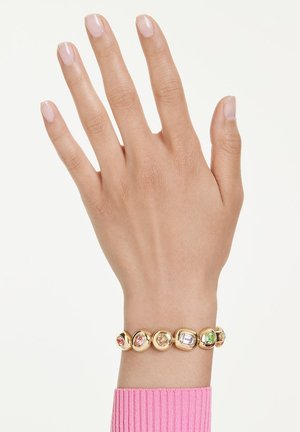 Χέρι με φυσικά νύχια που φοράει χρυσό βραχιόλι με στρογγυλές βάσεις και πολύχρωμους πολύτιμους λίθους, ροζ μανίκι ορατό στον καρπό.
