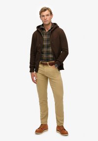 Hombre de pie wearing sudadera marrón, camisa de cuadros verde y naranja, pantalones beige, cinturón trenzado, botas de cuero marrón y reloj de pulsera negro.