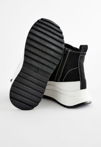 Stivaletto alla caviglia in pelle scamosciata nera con suola bianca, dotato di suola esterna in gomma nera rigata e cuciture bianche a contrasto lungo le cuciture.