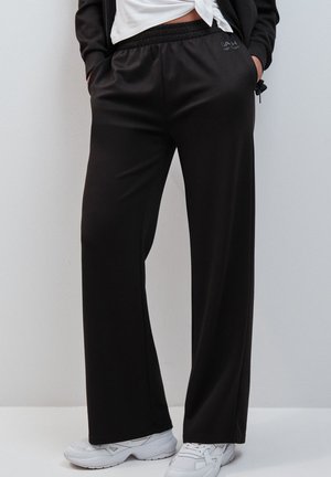 Personne portant un pantalon noir à jambes larges avec une taille élastique et des baskets blanches, debout les mains dans les poches devant un fond blanc.