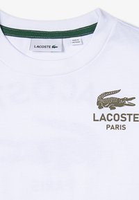 Biela tričko s logom zeleného krokodíla a textom "LACOSTE PARIS". Zelený lem pri golieri; štítky sú viditeľné pri výstrihu.