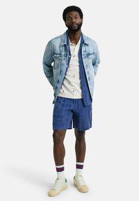 Denimjakke, hvid skjorte med sejlskibsdesign, blå shorts og cremefarvede sneakers med grønne accenter; struktureret stof og sporty design.