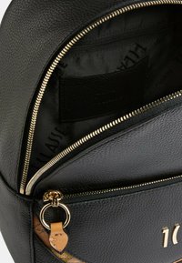Czarny plecak ze skóry o teksturowanej powierzchni, ze złotym suwakem i brązowym skórzanym akcentem. Zawiera wewnętrzną kieszeń oraz detale z logo.