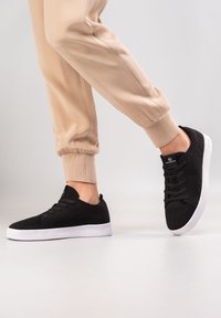 Czarne dzianinowe sneakersy z okrągłym noskiem, sznurowane z przodu i białą gumową podeszwą. W połączeniu z beżowymi spodniami dresowymi z elastycznymi mankietami.