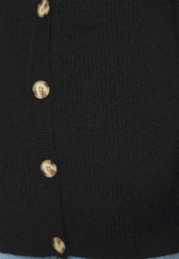 Närbild av en svart stickad cardigan med fyra stora beige marmoriserade knappar knäppta fram.