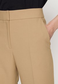 Beige högmidjade byxor med en slät yta, som har sidfickor och en skräddarsydd passform. Tyget verkar lätt och strukturerat.