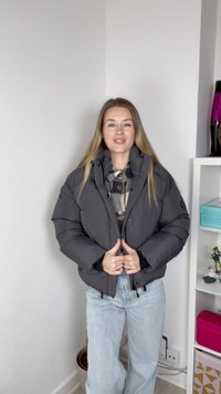 Casaco puffer cinza com colarinho alto, apresentando um design acolchoado e um enchimento espesso. Usado por cima de uma camisa xadrez e combinado com jeans de ganga clara.