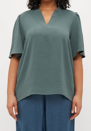 Femme portant un haut ample et texturé vert sauge à manches courtes avec un col en V et un pantalon bleu décontracté, debout devant un fond uni.