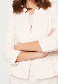 Veste rose clair avec un motif en losanges texturé, manches trois-quarts et design sans col, coupe structurée.