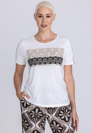Femme aux cheveux courts blonds portant un t-shirt blanc à motifs floraux et un pantalon assorti, debout avec une main dans la poche.
