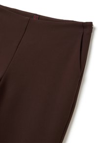 Gros plan sur un pantalon ajusté marron foncé montrant la ceinture, une poche latérale et une petite étiquette en tissu à l'intérieur de la ceinture.