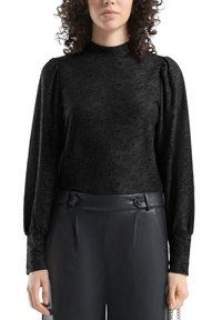 Czarna, teksturowana bluzka z długimi bufkami na rękawach i wysokim kołnierzem. Połączona z czarnymi spodniami z ekoskóry z wysokim stanem, ozdobionymi podwójnymi guzikami.