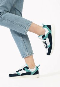 Marinblå och teal sneakers med övre del av mesh och mocka, vit sula och snördesign med en bakre hälflik. Bärs med avskurna jeans.
