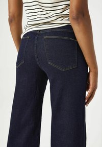 Jeans de mezclilla oscura de pierna ancha con dos bolsillos traseros, acentos de costura dorada y una cintura alta. Combinados con una blusa de rayas.