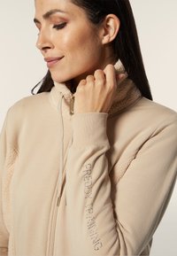 Giacca zip-up beige con colletto e polsini texturizzati e pelosi, con "FREDDY TRAINING" impreziosito da strass sulla manica.