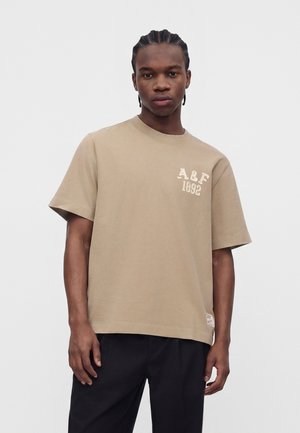 Junger Mann mit geflochtenem Haar, der ein beiges A&F 1892 T-Shirt und schwarze Hose trägt, steht vor einem schlichten weißen Hintergrund.